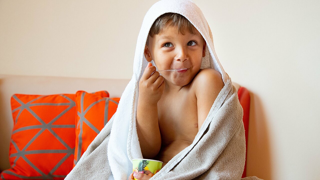 Frech und fröhlich lächelt ein Junge im Kapuzenbademantel während er ein Eis nach seiner Kindermassage genießt.